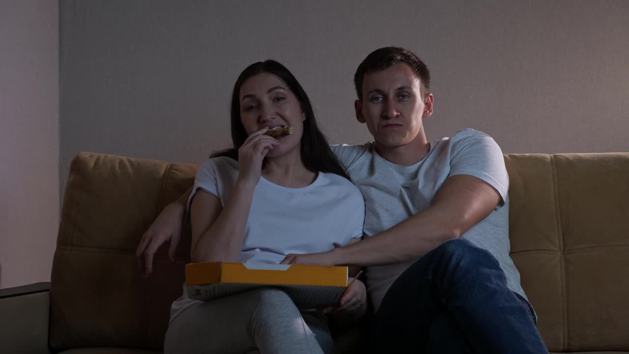 男人和女人坐在沙发上吃比萨饼和看电视视频素材