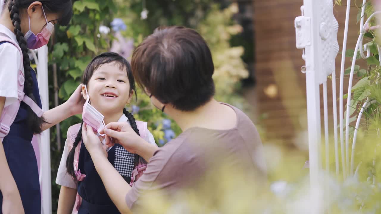 回到学校的亚洲华人母亲帮助她的女儿戴防护口罩从家到学校的前院早上视频素材