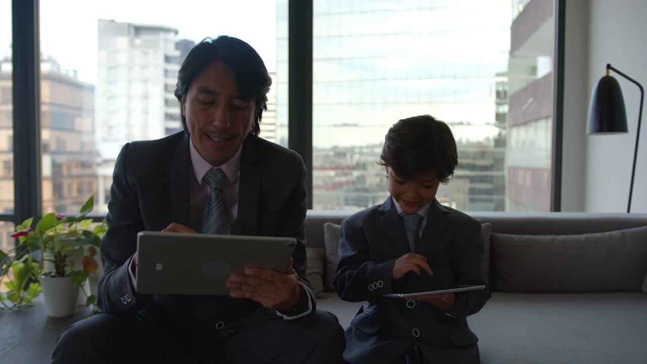 父子俩都穿着商务套装坐在沙发上玩着平板电脑视频下载