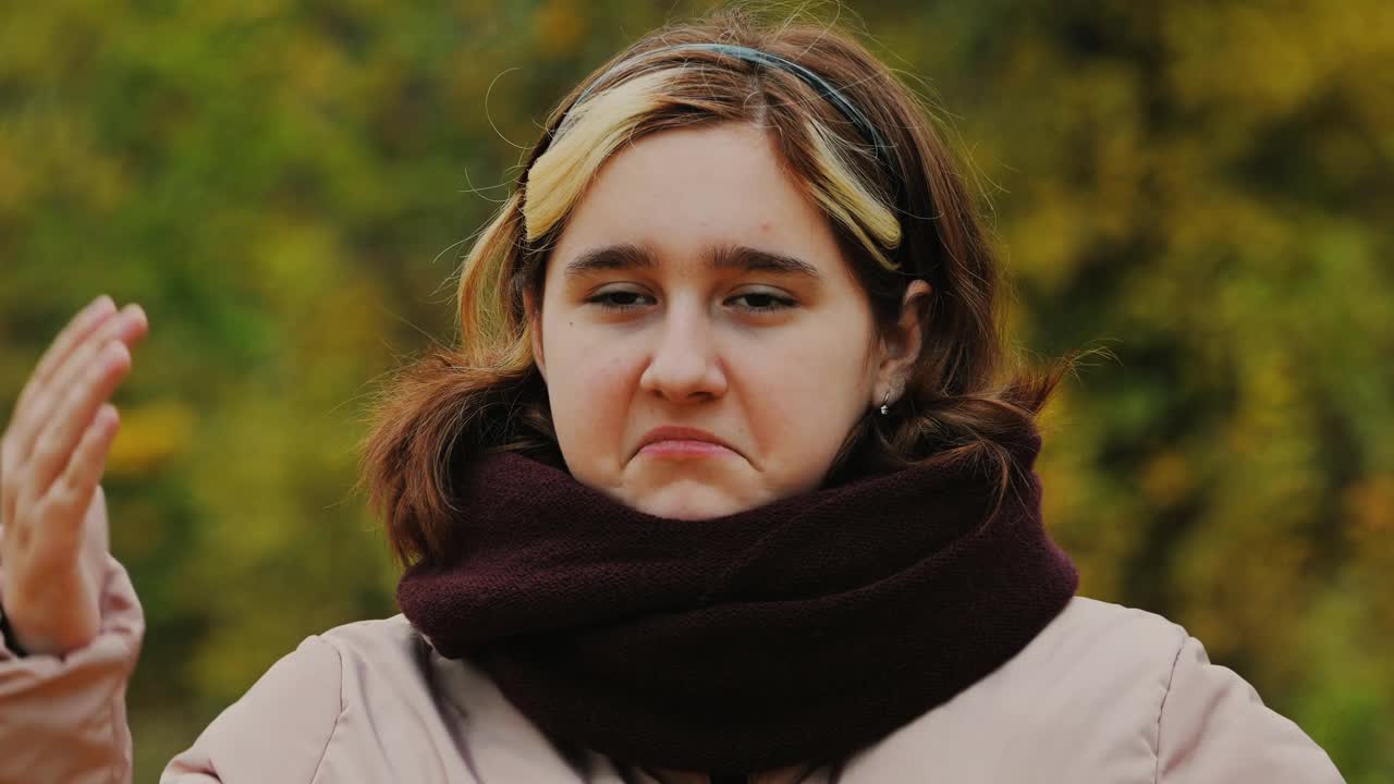 一个十几岁的女孩在秋天黄色的公园里做鬼脸，挥手在她的脸上。视频下载