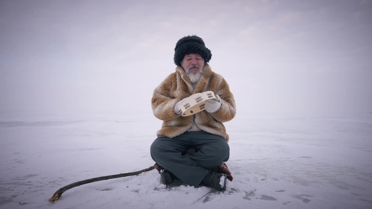广角镜头:老蒙古人坐在外面结冰的雪地上，用慢动作演奏着手鼓。前视图肖像集中自信的男子与极地自然在背景。视频下载