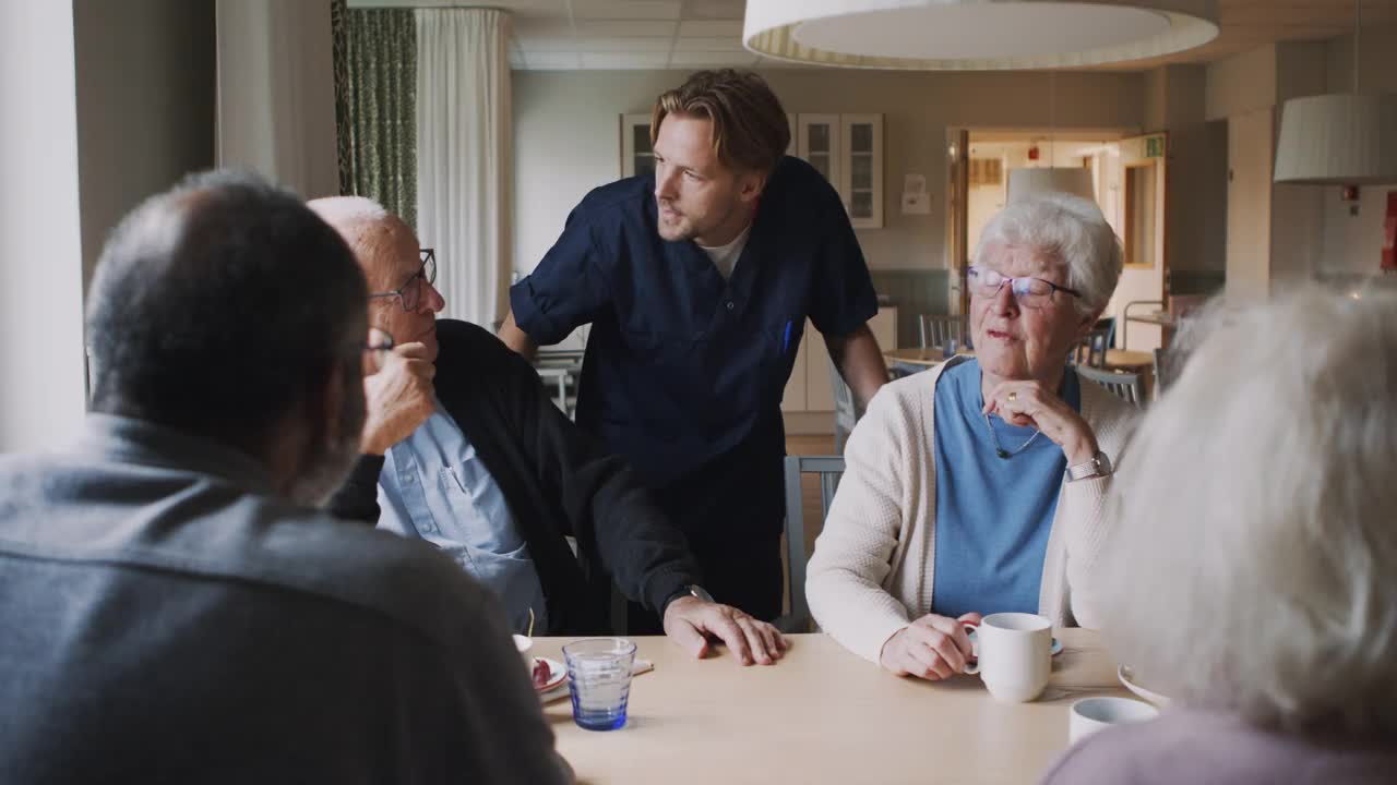 在敬老院，微笑的男护士和喝咖啡的老年男女交谈视频素材