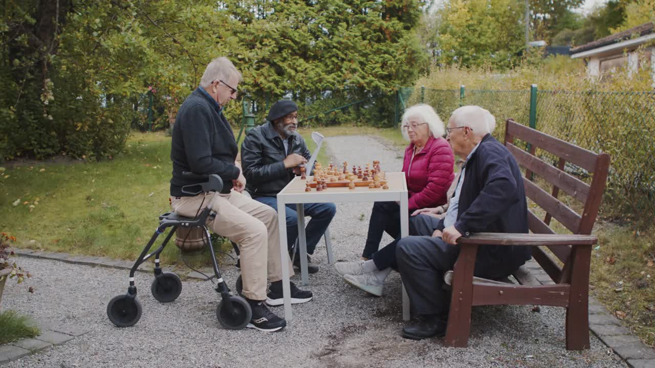 年长的男人和女人在下棋，而男性看护者在公园和他们交谈视频素材