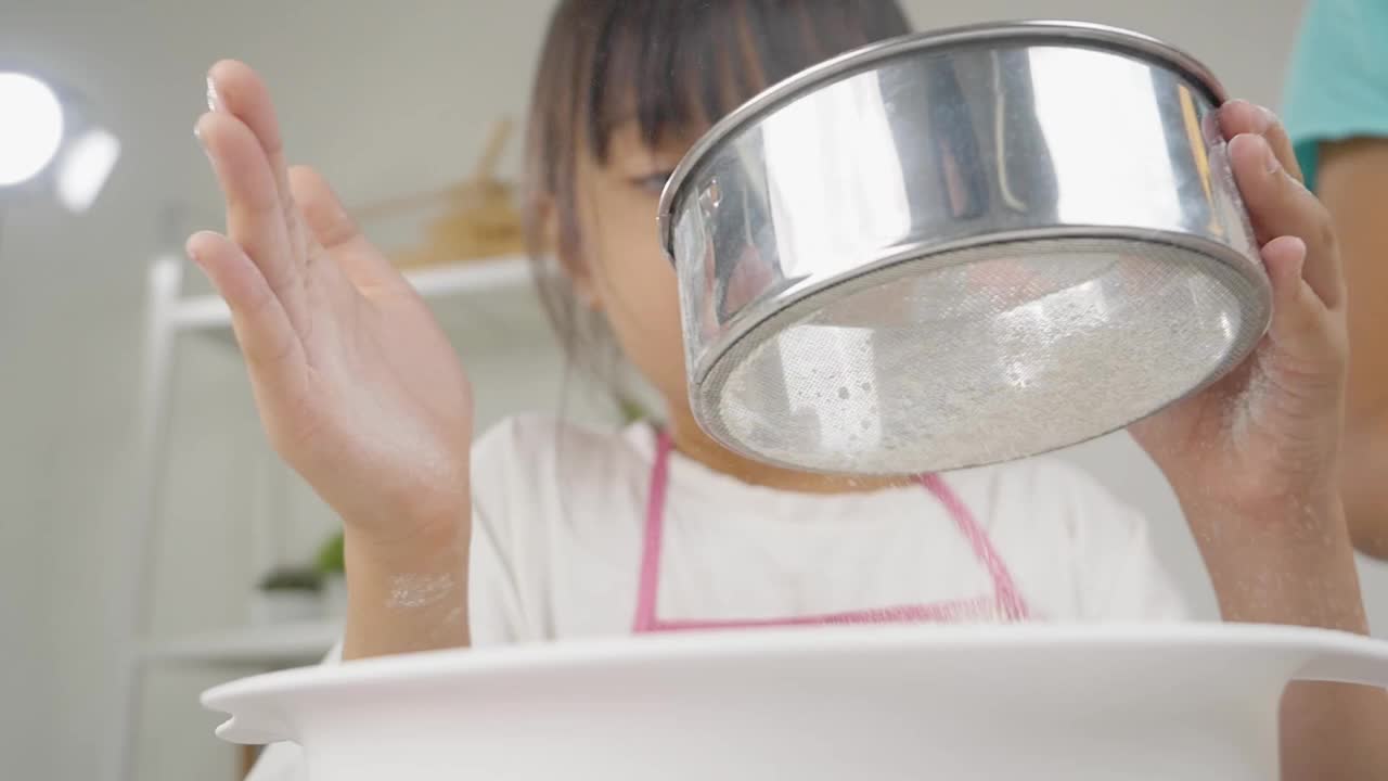 快乐的亚洲孩子在家里学习做饼干，女孩筛面粉到搅拌碗。视频素材