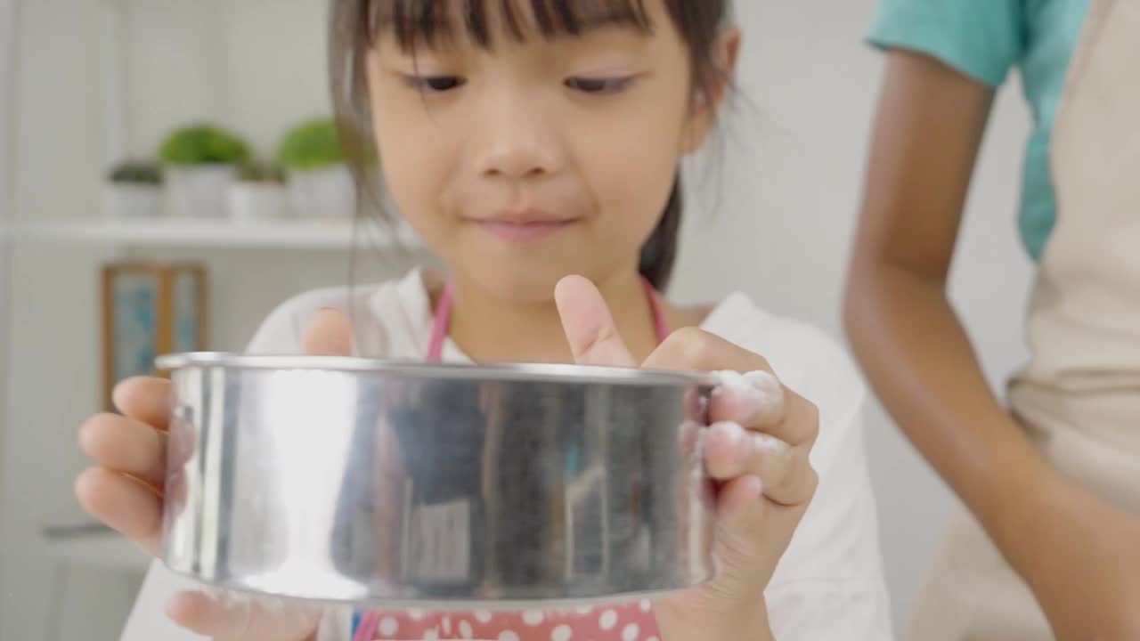 快乐的亚洲孩子在家里学习做饼干，女孩筛面粉到搅拌碗。视频下载