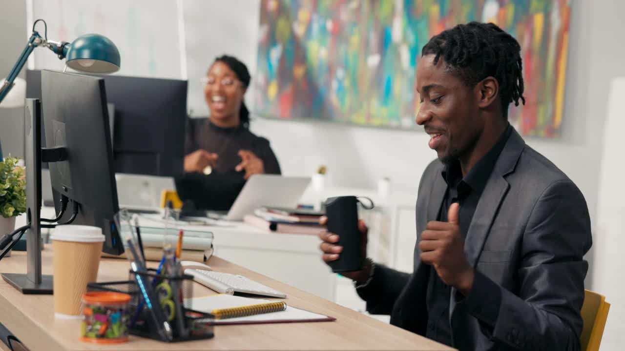 深色皮肤的同事坐在公司办公室的电脑前，跳舞，唱歌，为胜利而高兴，获得晋升，即将结束工作，良好的氛围，有趣视频素材