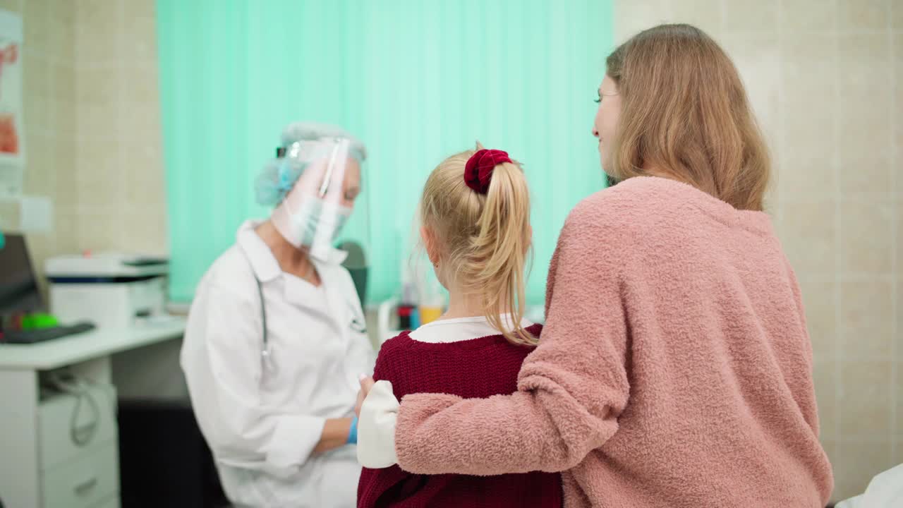放大架聚焦拍摄的高级女医生戴着防护面罩、面罩和白大褂与带着小女儿去看儿科医生的母亲交谈。妈妈耐心地拥抱孩子视频素材