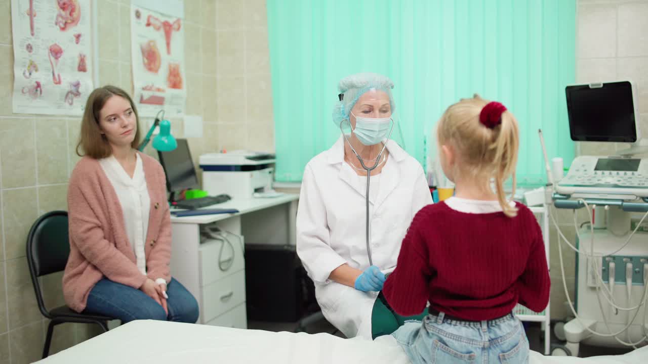 高级女儿科医师戴着防护面罩、面罩、白大褂，用听诊器聆听肺炎康复患儿的肺音，随母亲出诊视频素材