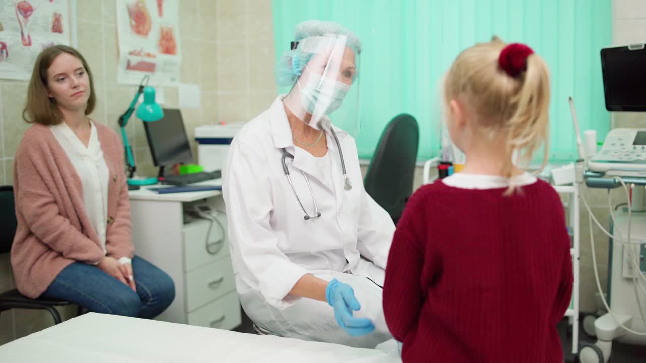 高级儿科医生戴着防护面罩、口罩和白大褂，为患儿检查，与关怀的母亲亲切交谈视频素材