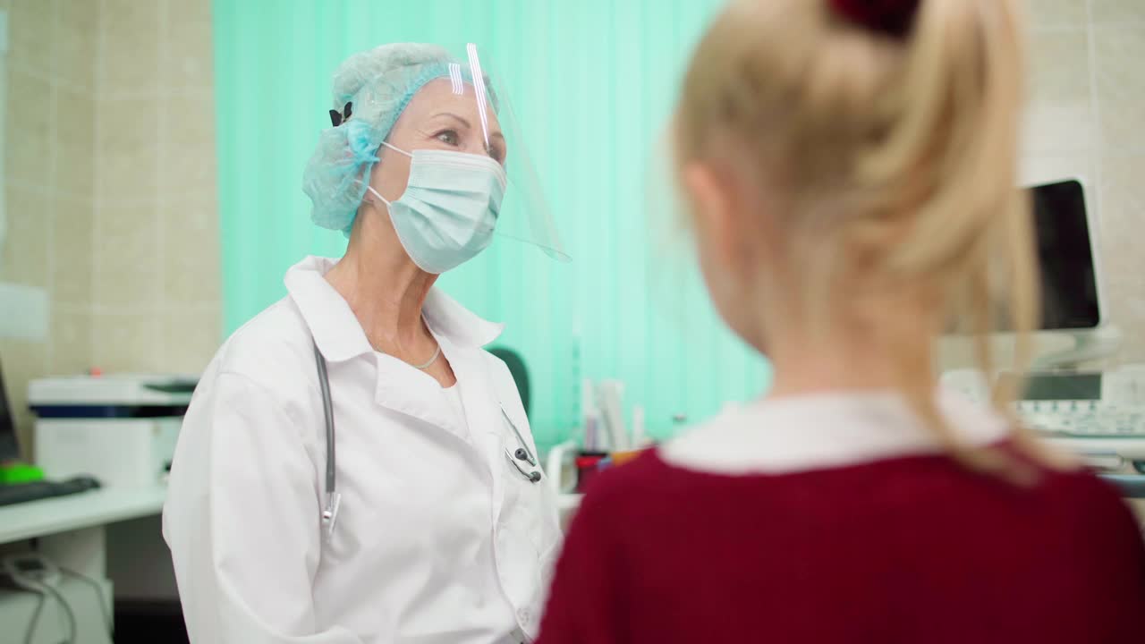 资深女医生戴着防护面罩和口罩与带着小女儿去看儿科医生的年轻母亲交谈。妈妈耐心地拥抱孩子视频素材
