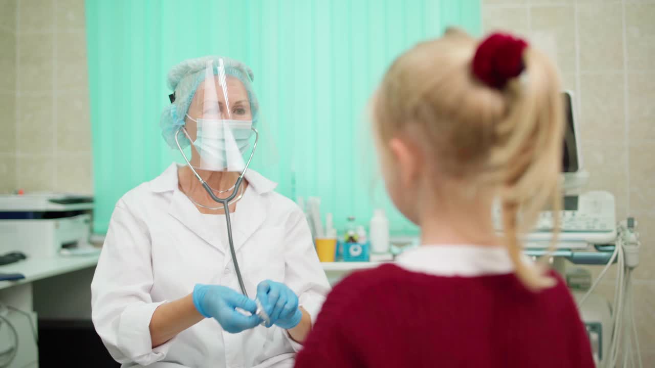 高级女医生戴着防护面罩、口罩、白大褂，用听诊器聆听肺炎、流感患儿的肺音。儿科医生与女孩友好交谈视频素材