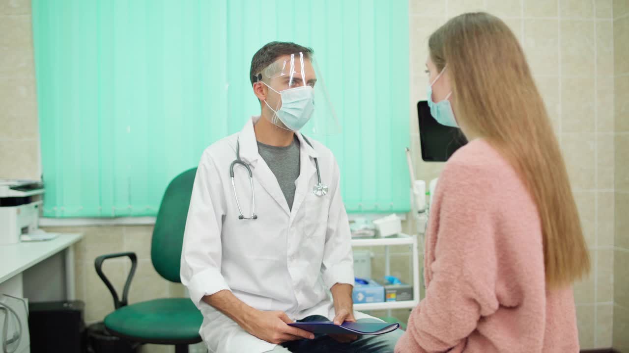 医生戴着防护面罩，手拿病历，在门诊与戴口罩的女患者交谈。身穿白大褂的年轻男性医生，负责治疗冠状病毒covid-19患者视频素材