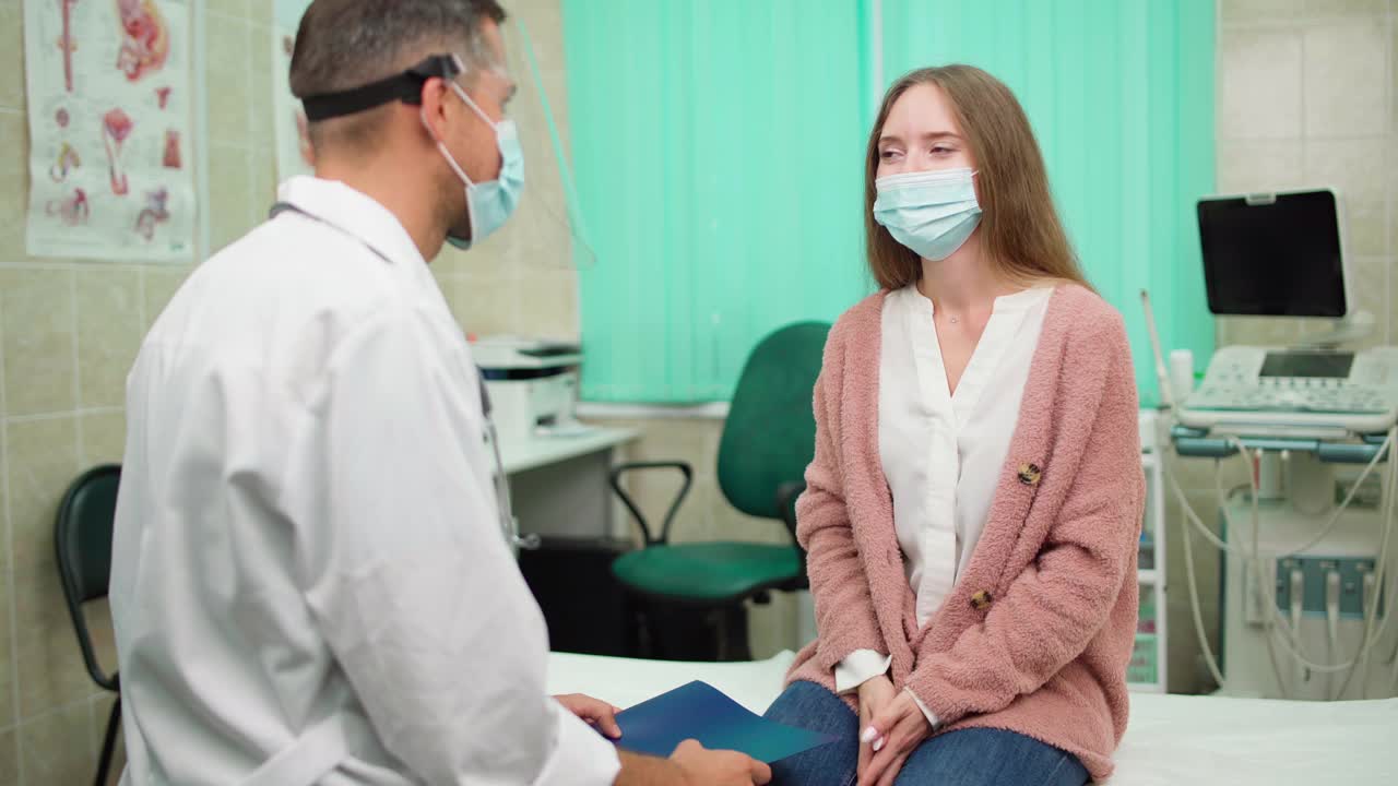 在医生办公室的沙发上，戴着面罩的医生与戴着面罩的年轻女病人交谈，并咳嗽。一名妇女在covid-19诊所解释她的疾病症状视频素材