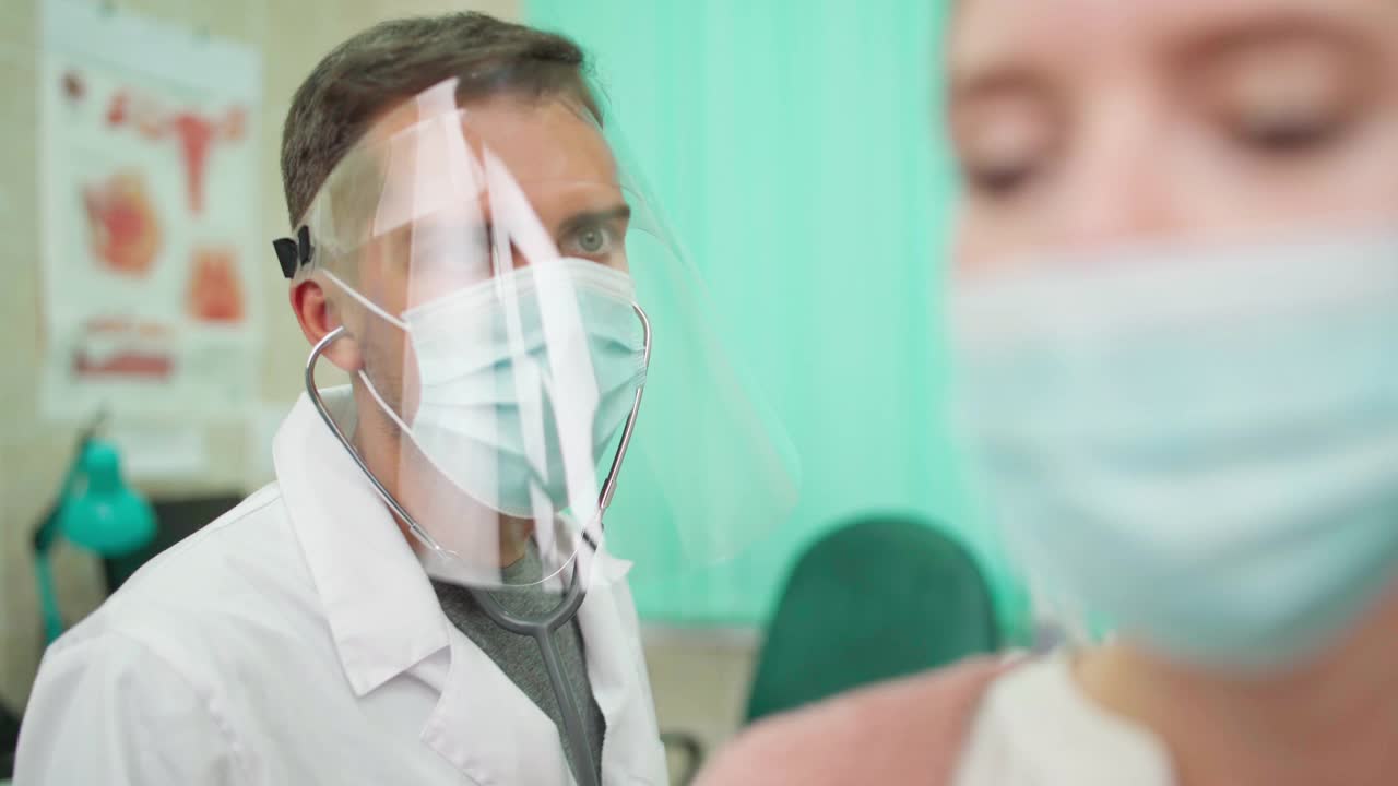 医生在防护面罩上用听诊器聆听年轻女性患者的肺音，戴口罩咳嗽，患冠状病毒covid-19肺炎视频素材