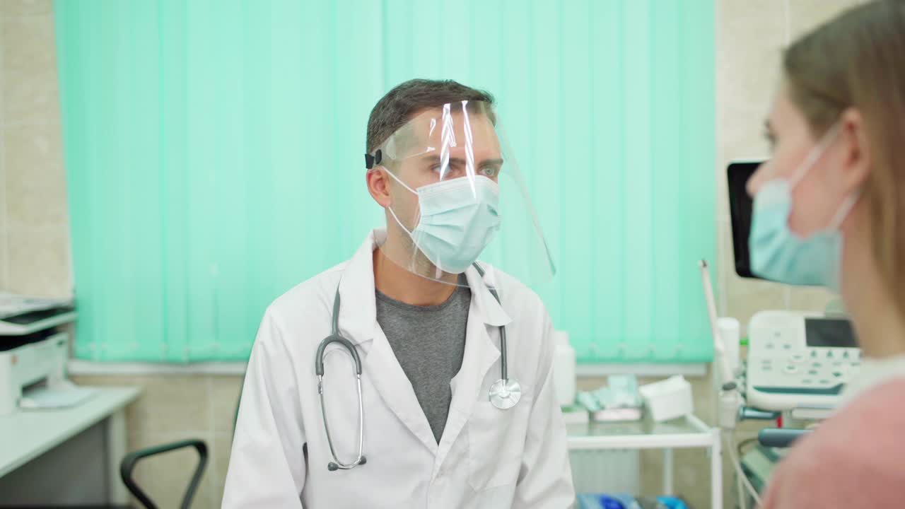 在门诊中，医生戴着防护面罩与戴着口罩的女患者交谈。身穿白大褂的年轻男性医生，负责治疗冠状病毒covid-19患者。医疗保健概念视频素材