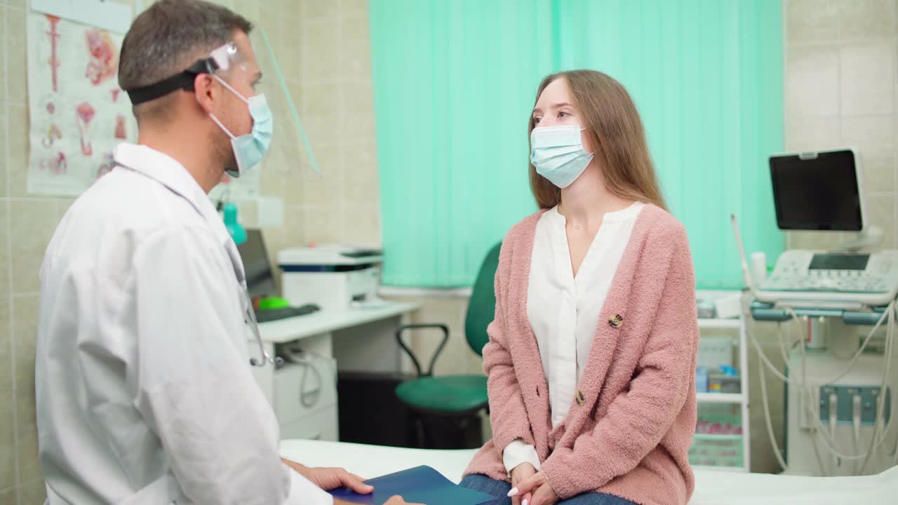 在医生办公室的沙发上，戴着面罩的医生正和戴着面罩的女病人交谈。妇女在诊所向医生解释她的疾病症状视频素材