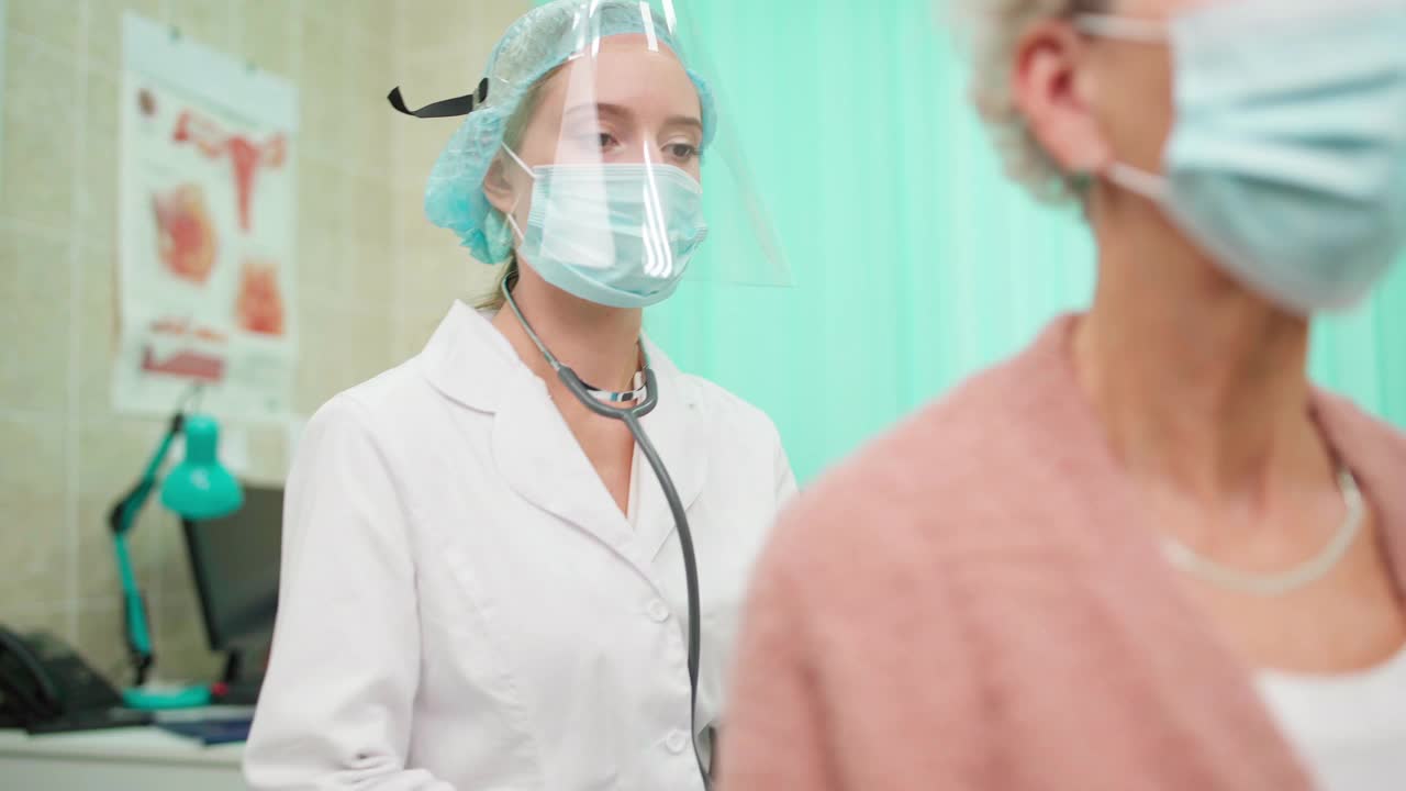 画面中，医生在防护面罩下用听诊器听着戴面罩深呼吸的老年女病人的肺音。冠状病毒大流行的概念视频素材