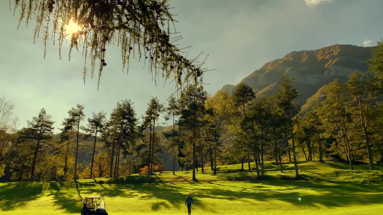 阳光明媚的一天，驾着高尔夫球车的高尔夫球手在有树和山的高尔夫球场用高尔夫球杆击球视频素材