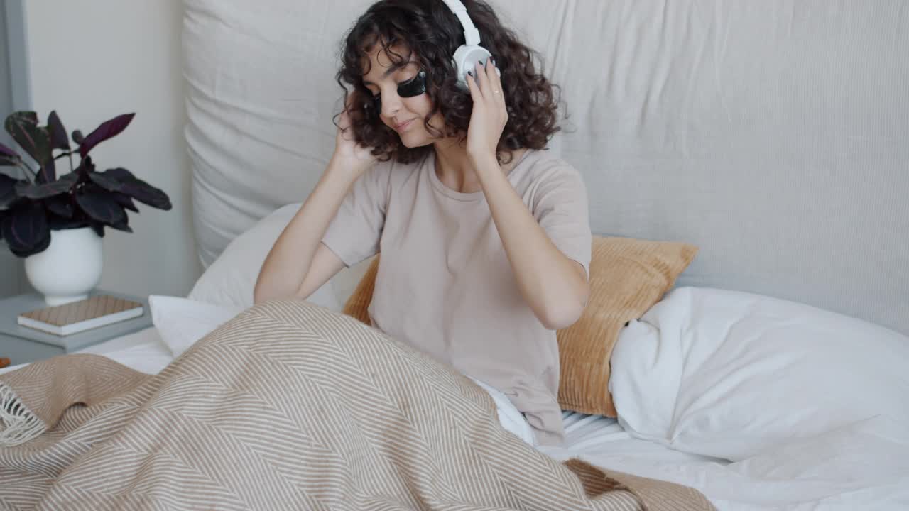 戴着眼罩的快乐女孩戴着耳机听着音乐在床上跳舞视频下载