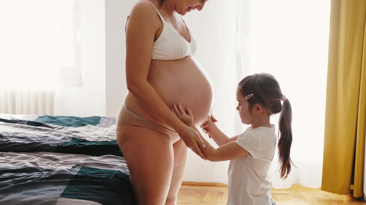 怀孕的中年妇女和家里的女儿。女孩亲吻怀孕的腹部视频下载
