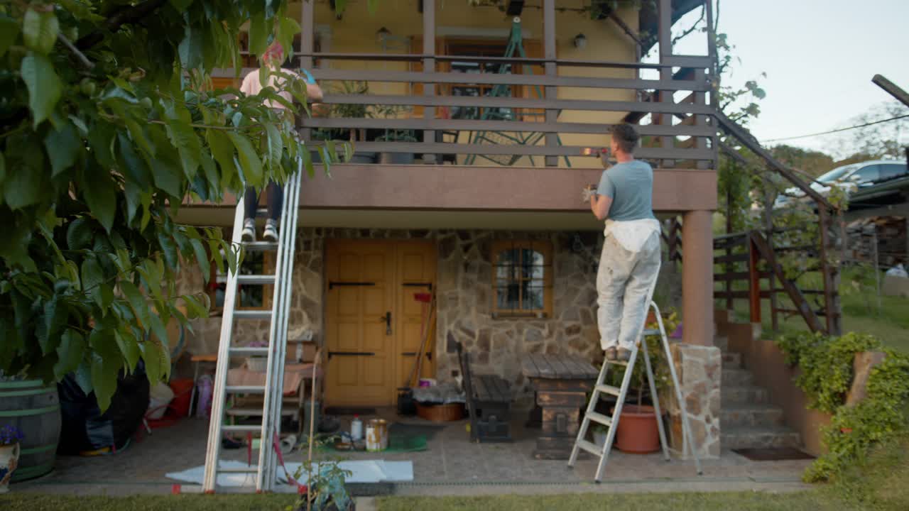 SLO MO夫妇用梯子粉刷阳台上的木栅栏视频素材