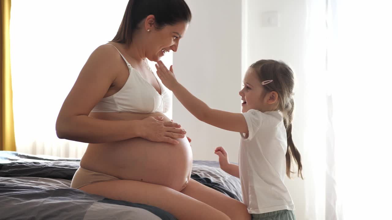 怀孕的中年妇女和家里的女儿。女孩在妈妈身上涂润肤霜视频下载