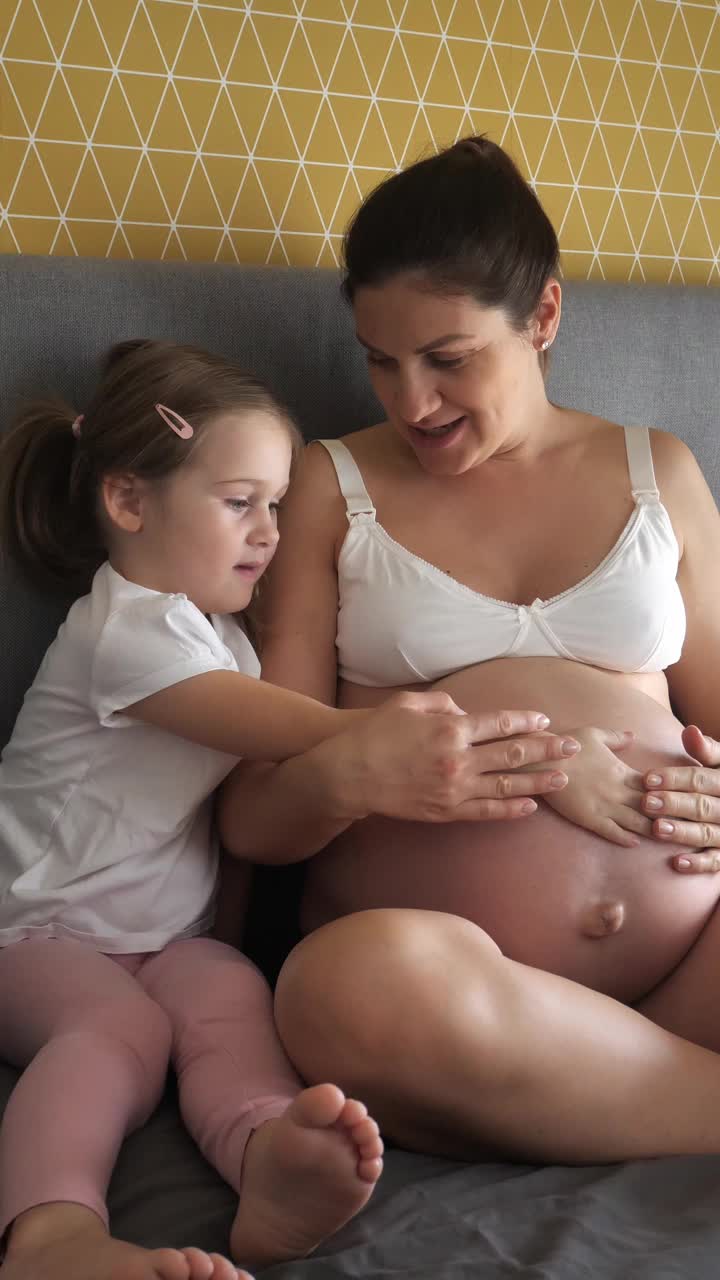 怀孕的中年妇女和家里的女儿。女孩抚摸孕妇腹部视频下载