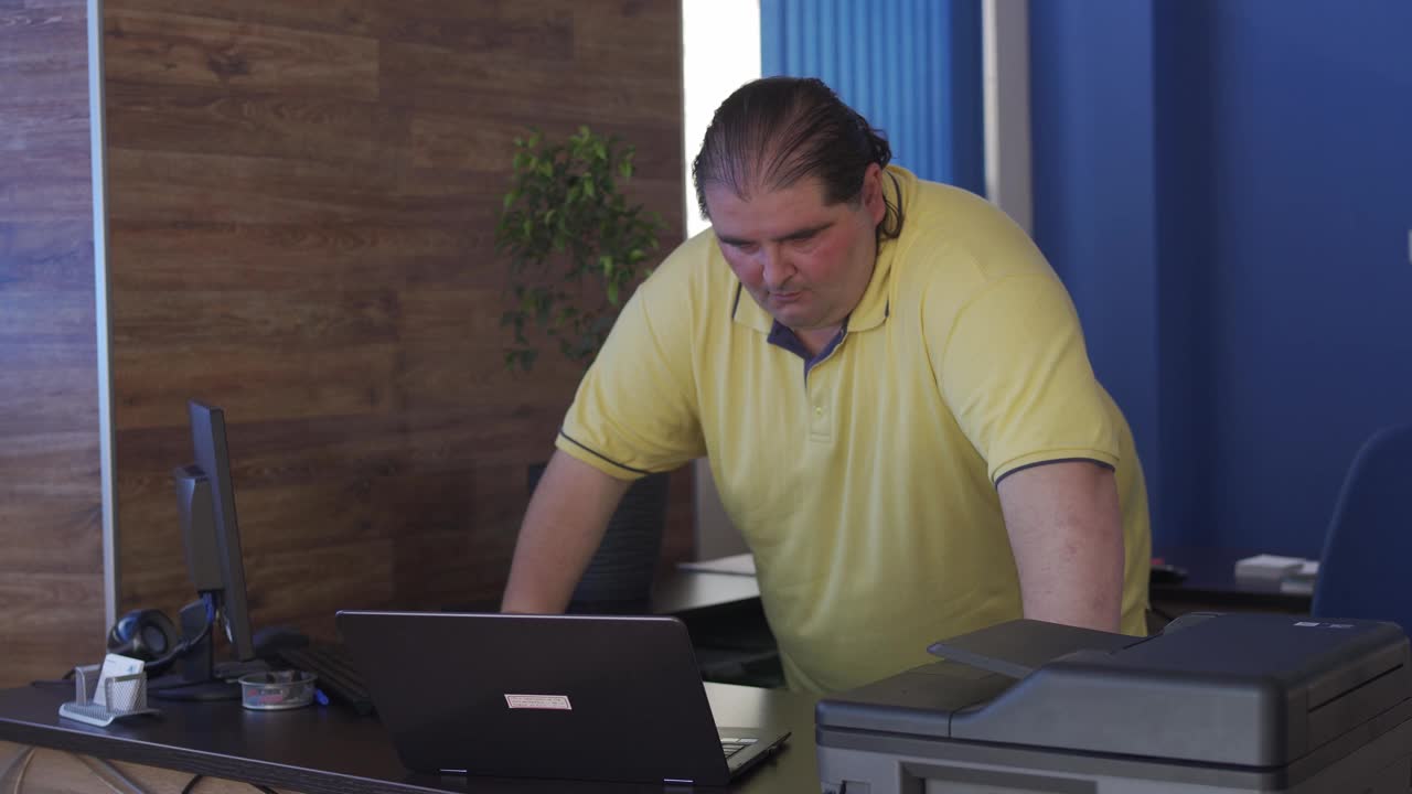 一个身材高大的男人倚在他的桌子上，看着他的笔记本电脑视频下载