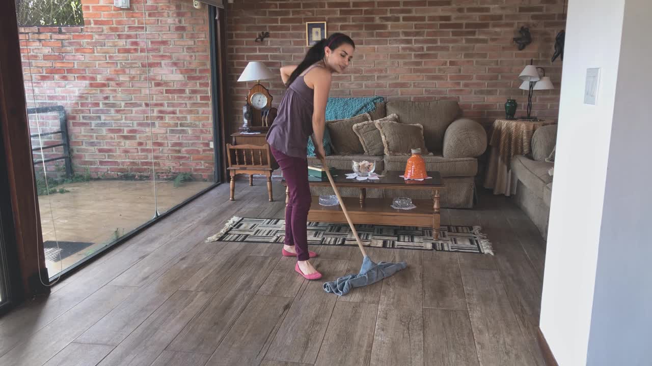 西班牙年轻女子一边跳舞一边清洁客厅的硬木地板和一个装清洁用品的塑料桶视频素材