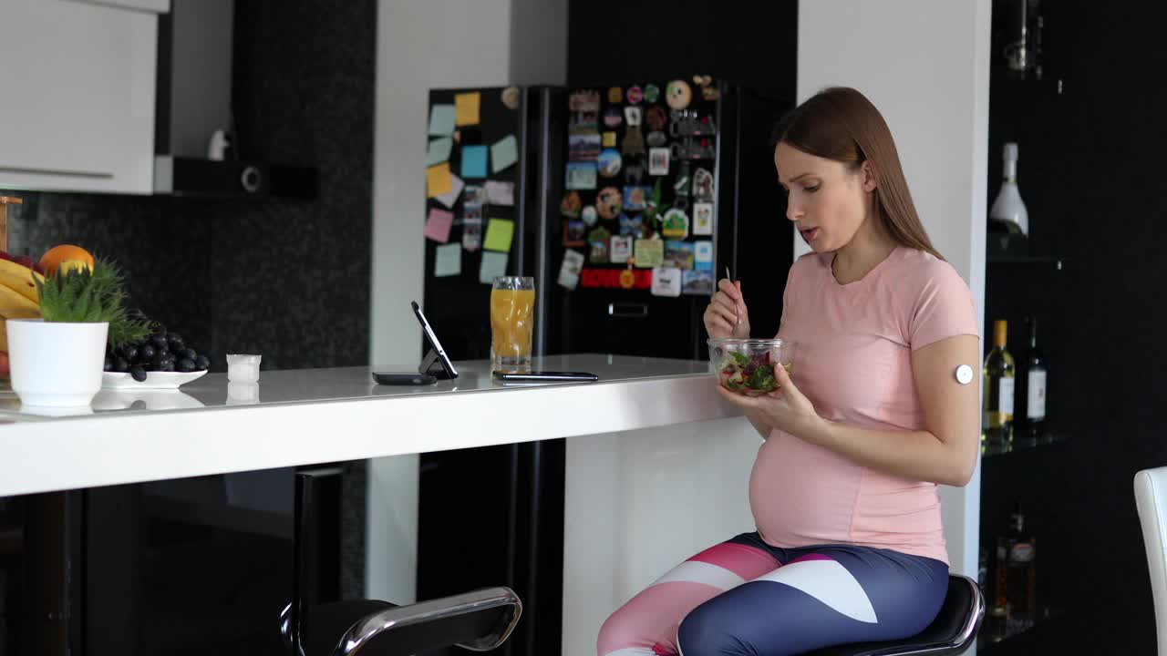 一位美丽的孕妇坐在厨房的吧台前，一边用平板电脑看电视节目，一边吃着碗里的健康沙拉。视频下载
