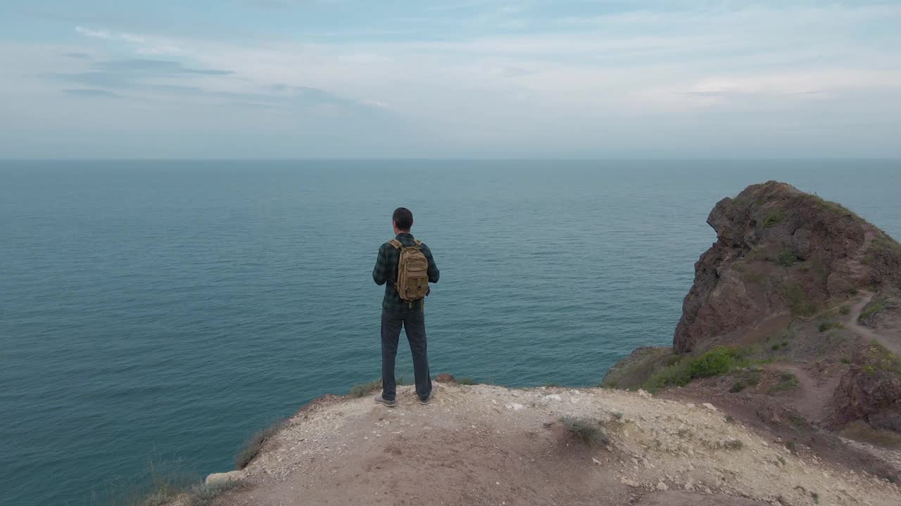 在户外休闲娱乐活动中，活跃的徒步旅行男子背着背包站在蓝海与天空地平线附近的岩石山上，航拍无人机视图视频下载