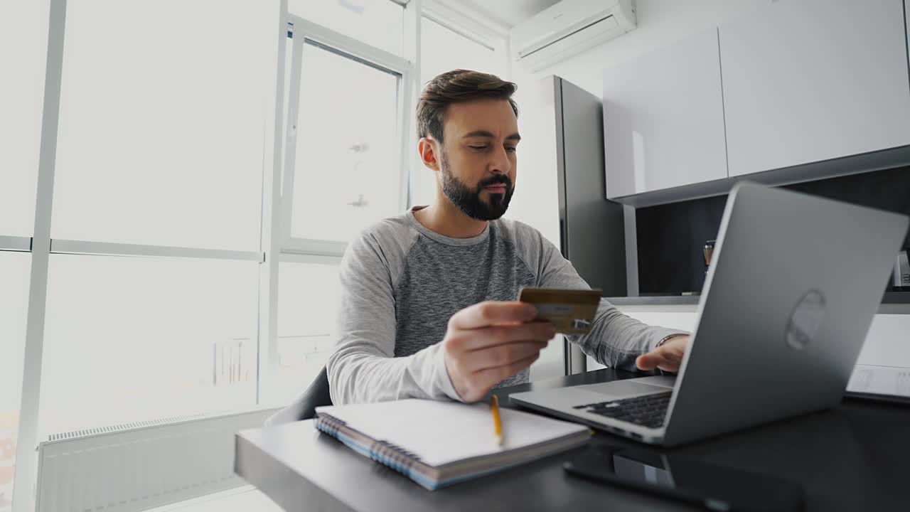 满脸胡须的微笑男子一边用笔记本电脑工作，一边用信用卡付款视频素材