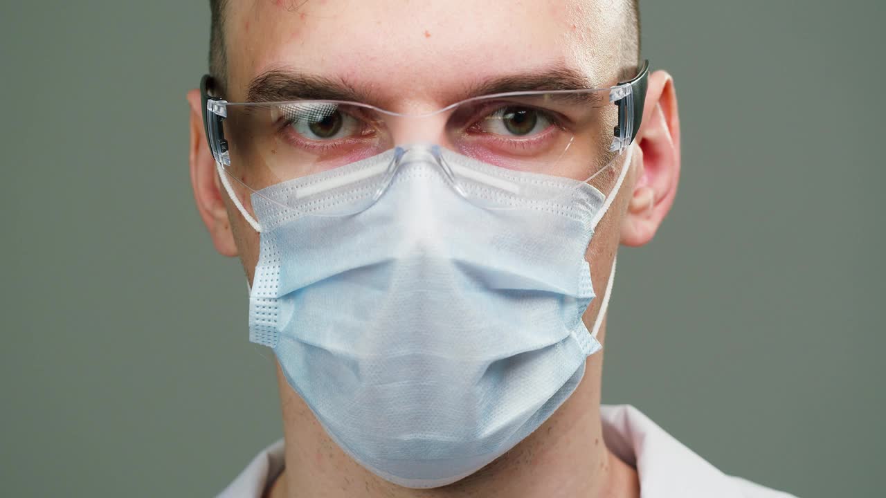 特写的医生，科学家专家戴着医用防护眼镜和面具，男性专业化学家的肖像在制服看着相机，孤立在灰色背景视频下载