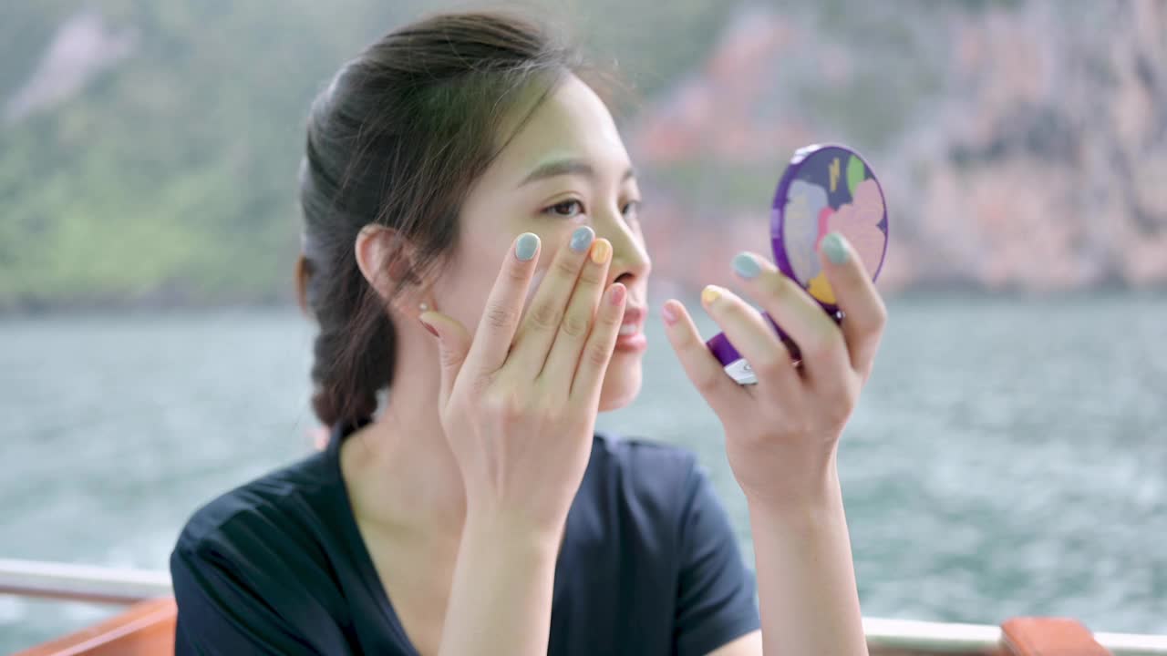 在热带海滩上，一名亚洲泛亚妇女正在给自己的脸上涂防晒霜，在享受夏天之前，她在船上微笑视频下载