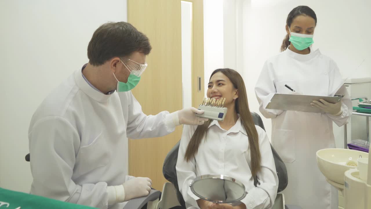 一名白人牙医，在医院诊所，人们比较牙齿贴面图与亚洲妇女的牙齿。牙齿美白。医生。牙医。医疗和牙科健康。视频下载