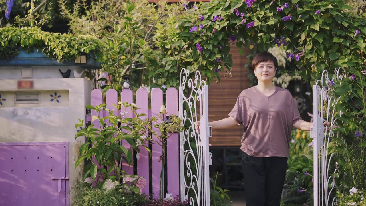 在清晨的阳光下，亚洲华人中年妇女站在充满牵牛花的房子的前院看着摄像机视频下载