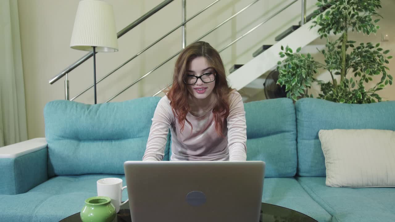 戴着眼镜微笑的美丽女孩在电脑前工作，坐在家里的沙发上享受着好消息视频素材