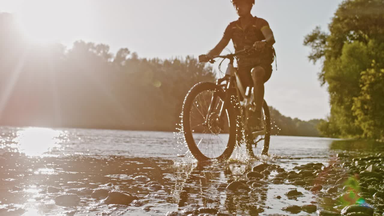 日落时分，山地车手在河的浅水中骑自行车的时间扭曲效应视频下载
