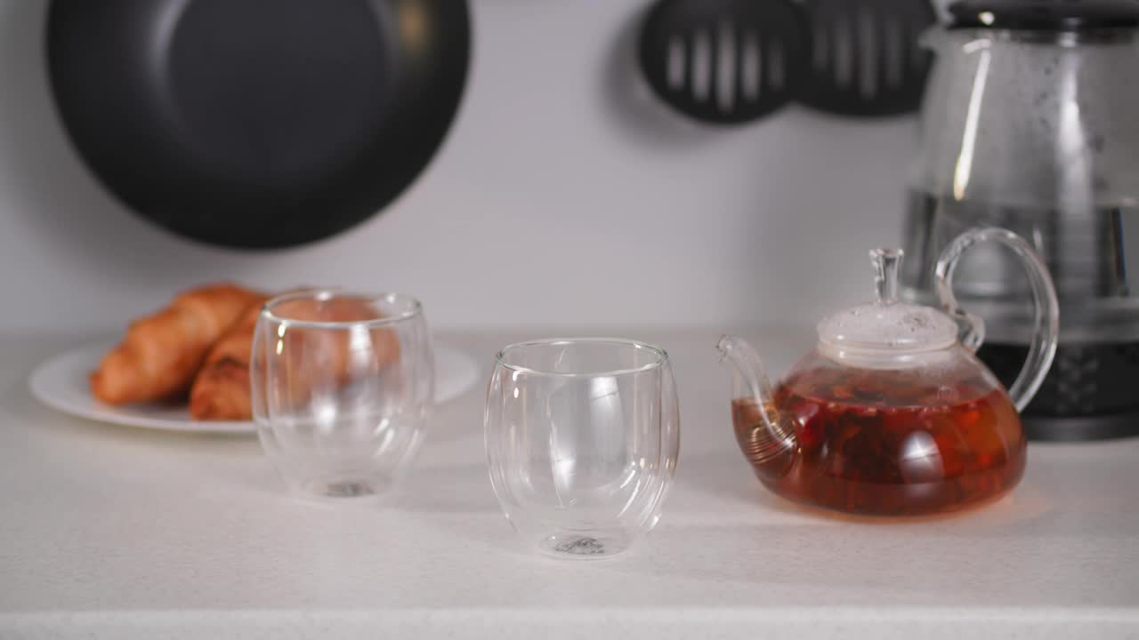 喝茶的时候，女人用手把带花的热饮从透明水壶里倒进玻璃杯子里，背景是放在盘子里的羊角面包视频素材