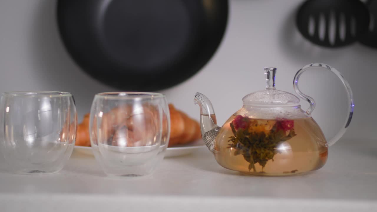 在厨房桌子上的透明水壶里，站在杯子旁边，以牛角面包为背景，泡茶的过程，早餐时间视频素材