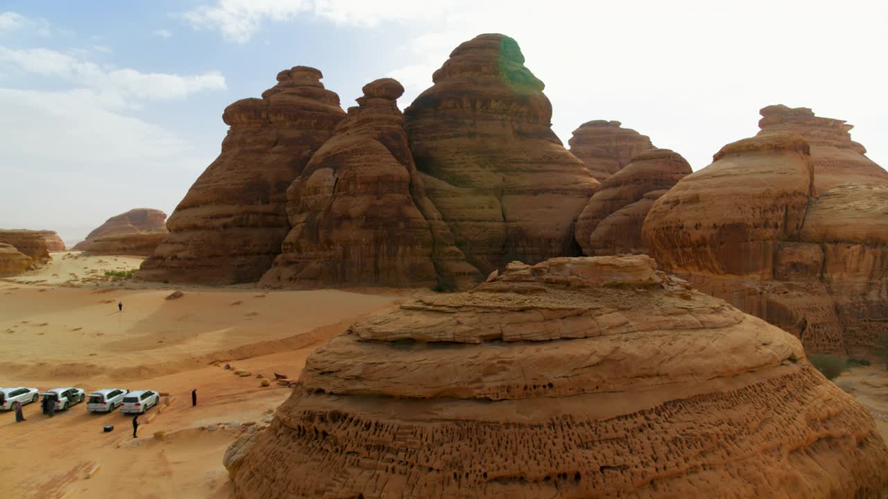 空中倒车远离一个大的沙漠岩层和古老的坟墓与汽车停在附近-赫格拉，沙特阿拉伯视频下载