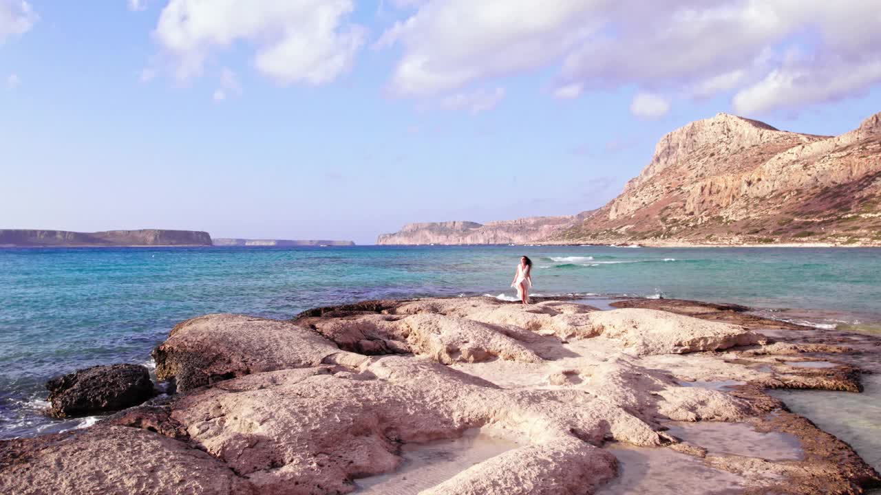 在阳光明媚的暑假里，一位身穿白色太阳裙、留着黑色长发的美女在美丽的海滩上的岩石上漫步。视频下载
