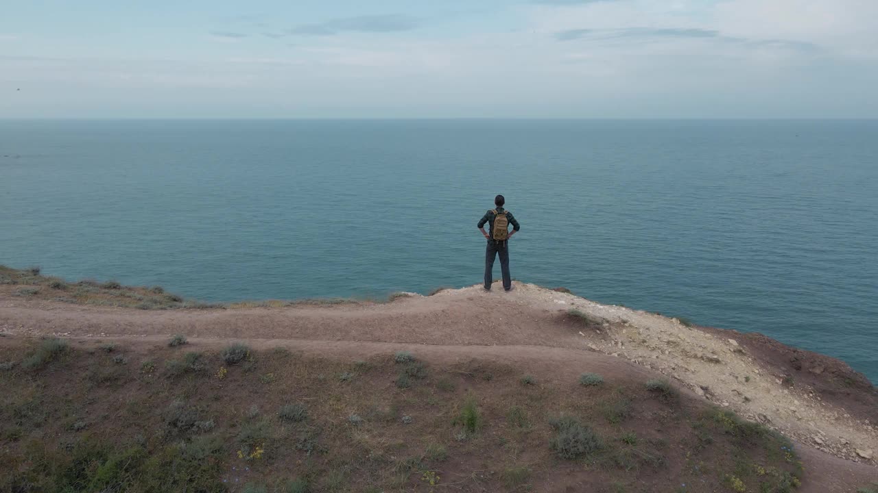 在户外休闲娱乐活动中，活跃的徒步旅行男子背着背包站在蓝海与天空地平线附近的岩石山上，航拍无人机视图视频下载