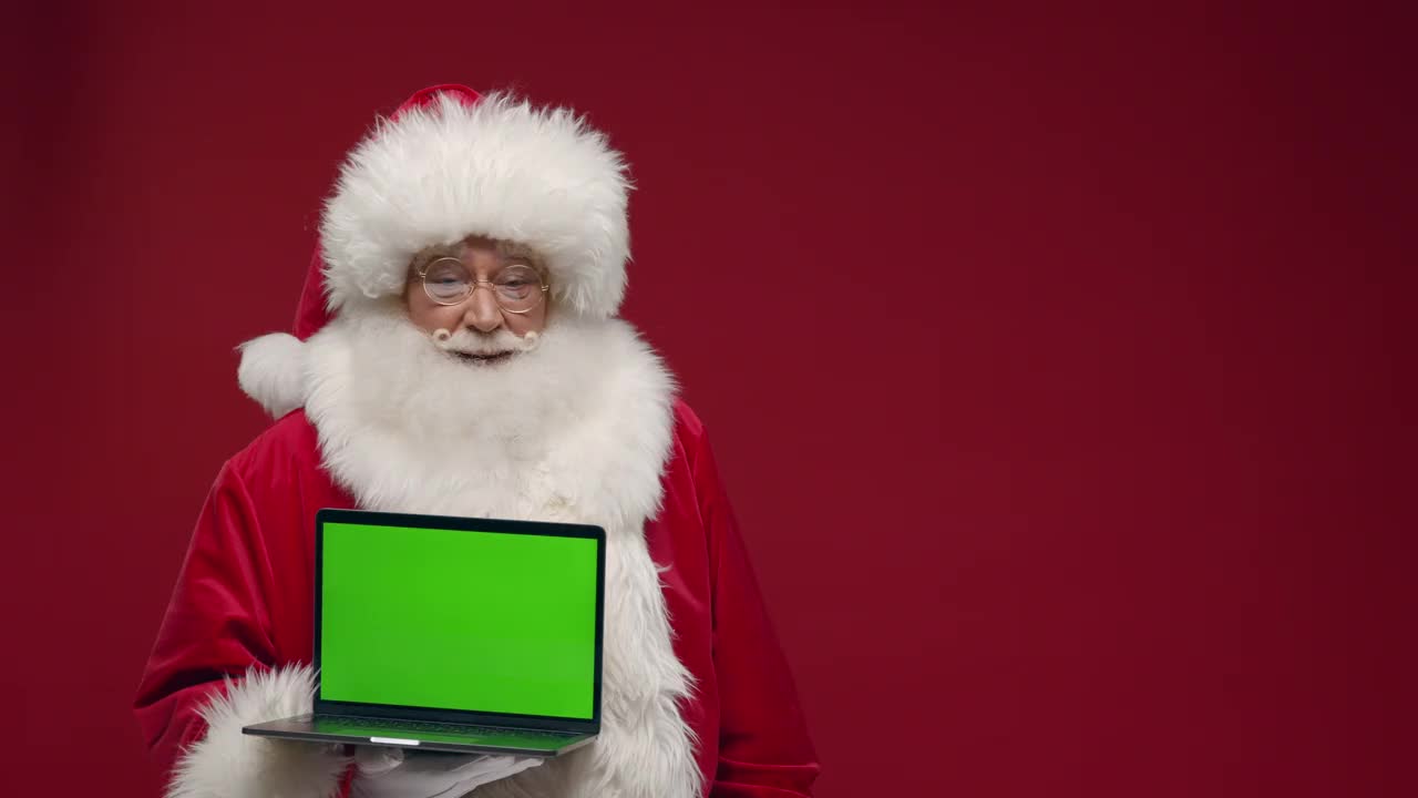 一位兴高采烈的圣诞老人站在红色的背景上，在相机里展示一个空的笔记本电脑的绿色屏幕，微笑着竖起大拇指视频下载