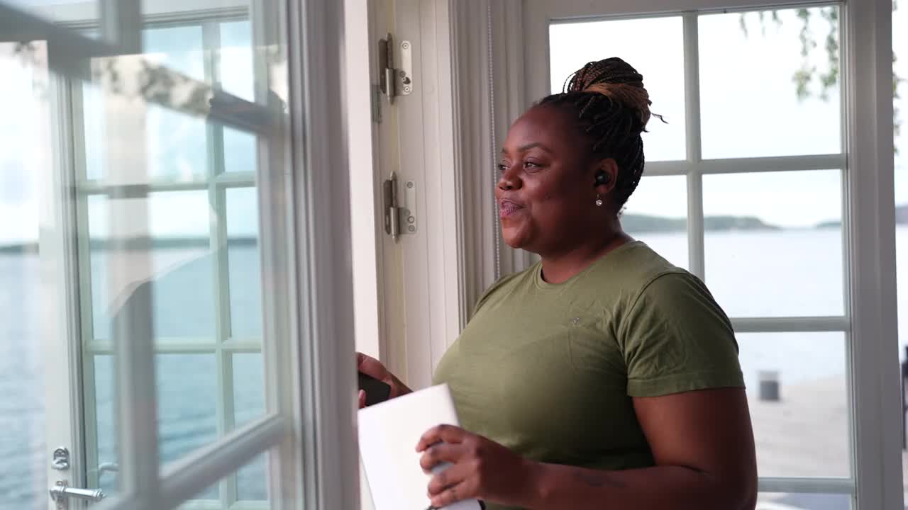 女自由作家，带着笔记本，站在小屋的窗户边用智能手机聊天视频下载