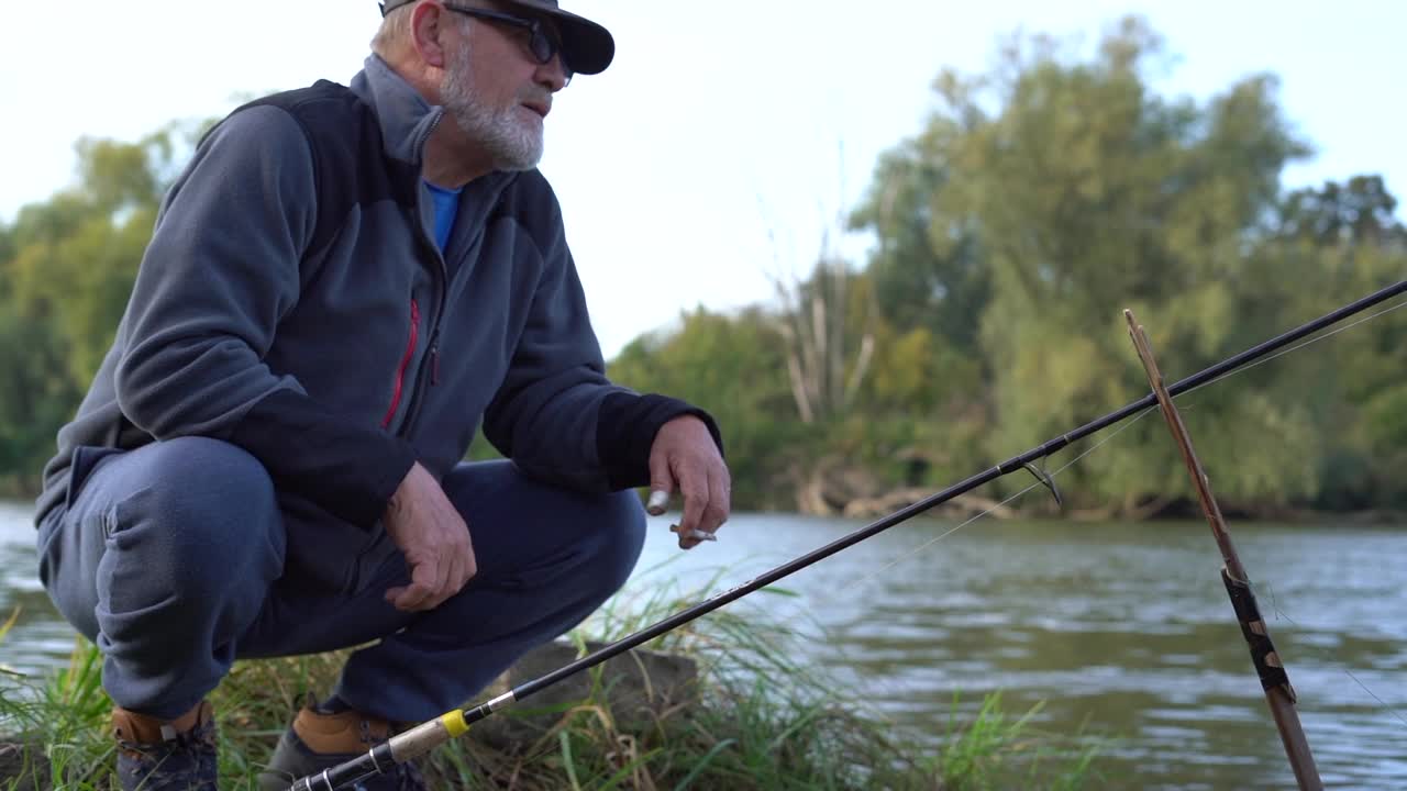 一位戴着帽子、戴着眼镜、嘴里叼着香烟的老渔夫坐在湖边的码头上，手里拿着鱼竿垂钓。视频素材