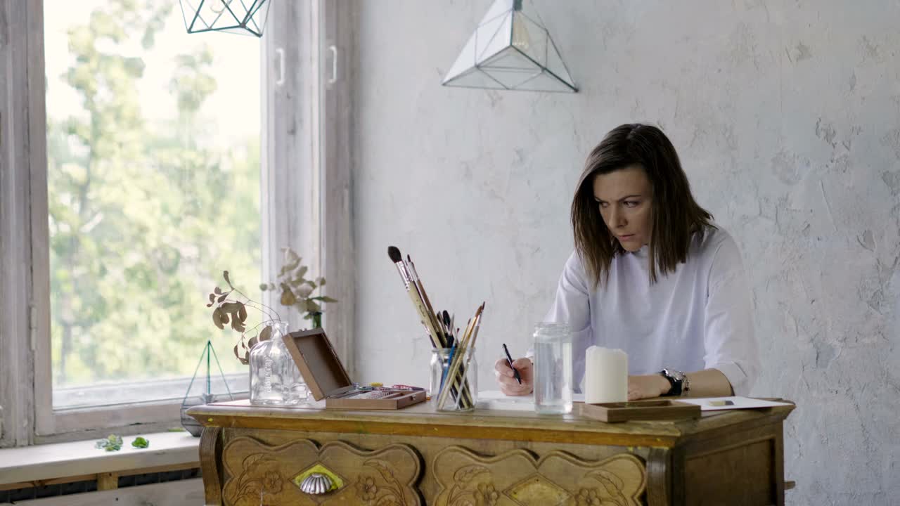 一个漂亮的黑皮肤女艺术家在木桌上用铅笔在纸上画画。4 k视频下载