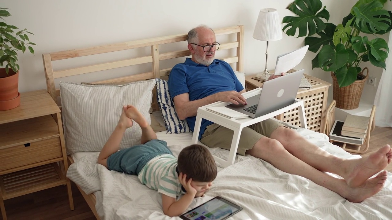 爷爷在笔记本电脑上工作，孙子在数码平板电脑上看动画片视频素材