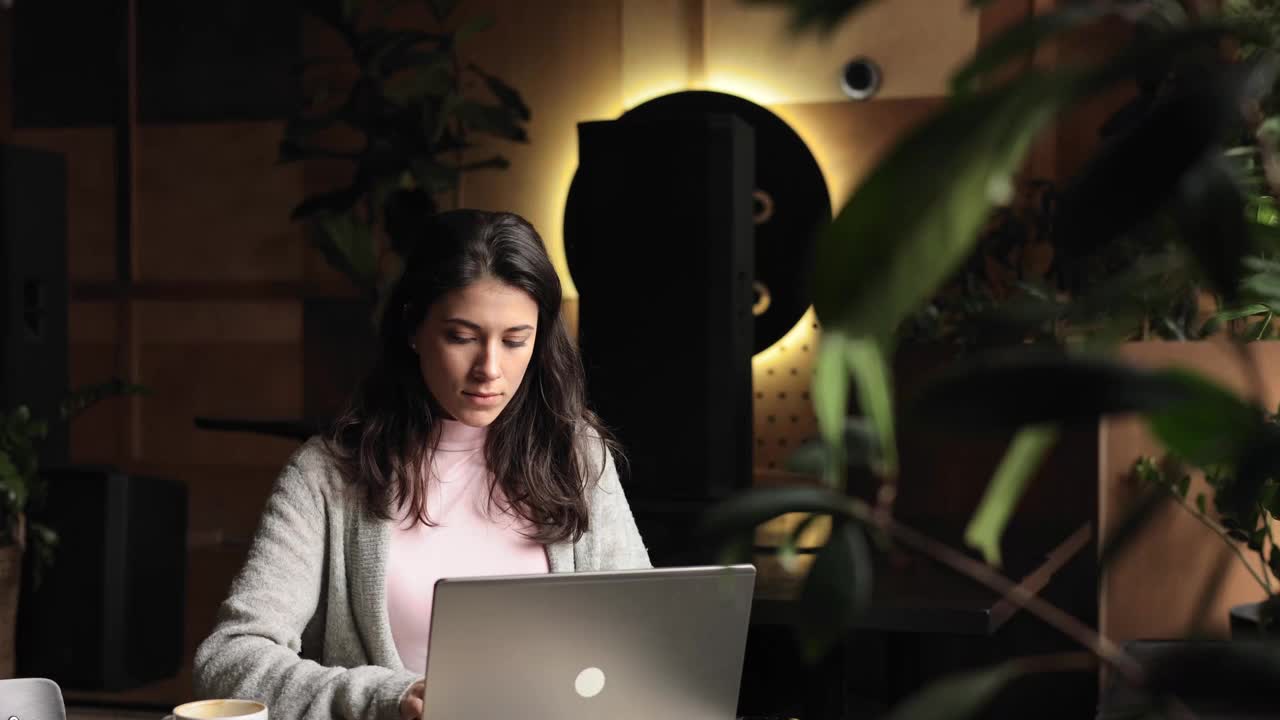 女人在笔记本电脑上打字，看着窗户思考。自由职业的黑发女性工作在咖啡馆坐着聊天。开朗的女员工，用笔记本电脑工作。视频下载