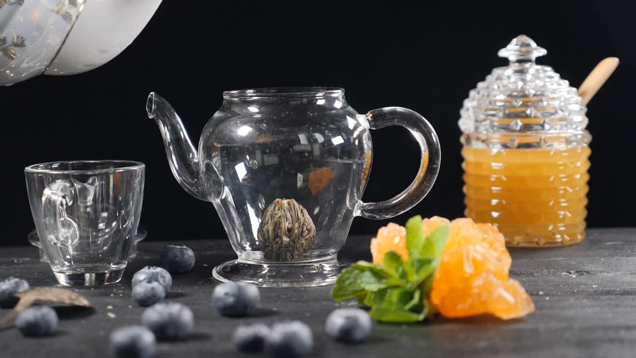 茶酿造。绿色中国茶花玻璃茶壶在木制背景。蓝莓，玻璃罐和蜂蜜。倒开水。缓慢的运动。在高清拍摄视频素材